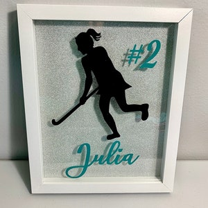 Peut inclure: Une boîte à ombre encadrée de blanc avec un fond de paillettes turquoise. Une silhouette noire d'une joueuse de hockey sur gazon avec un bâton est au centre. Le numéro "#2" est en turquoise au-dessus de la joueuse. Le nom "Julia" est en turquoise en dessous de la joueuse.