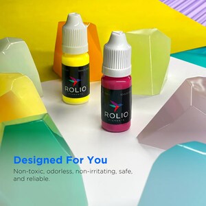May include: Two small bottles of paint, one yellow and one pink, with the brand name "ROLIO PIGMENTS" printed on the labels. The bottles are sitting on a white surface with colorful geometric shapes.