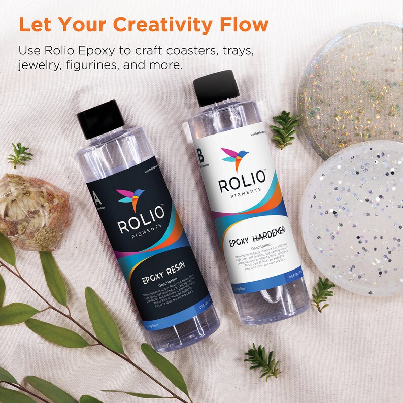 May include: Two clear bottles of Rolio Pigments epoxy resin and hardener. The bottles are labeled with the product name and description. The bottles are sitting on a white surface with a small green plant and two white coasters.