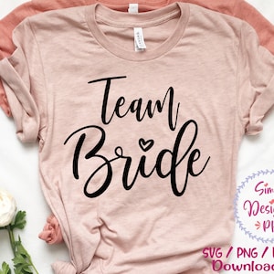 May include: A pink t-shirt with the text "Team Bride" in black script with a small heart above the word "Bride". The text is centred on the shirt. The shirt is folded and laid flat on a white surface.