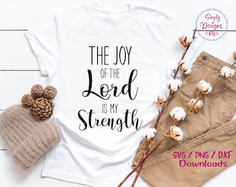 May include: White t-shirt with black text that reads "The Joy of the Lord is My Strength". The shirt is laying on a white wooden surface with brown pants, a brown knitted hat, and a branch with white cotton balls.