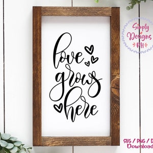 May include: A white sign with a brown wooden frame, featuring the text "love grows here" in black cursive lettering with small hearts surrounding the words.