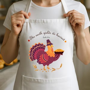 Op de afbeelding: Witte schort met een Thanksgiving kalkoen illustratie. De kalkoen draagt een pelgrimshoed en houdt een taart vast. Tekst op de schort luidt "The earth yields its harvest" en "Psalm 67:6".