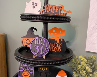 May include: A three-tiered black tiered tray decorated with Halloween-themed cutouts. The top tier features a ghost, a bat, and the words "Happy Halloween". The middle tier features a witch, a cauldron, and a purple circle with the text "Oct 31" and a spider web. The bottom tier features a black cat, a candy corn, a spider web, and the words "Boo".