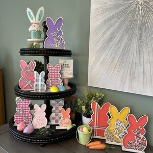 May include: A three-tiered black tiered tray with a variety of colorful Easter bunny decorations. The bunnies are made of wood and have different patterns and designs. There are also some small green plants and a small green bucket with plastic eggs on the tray.