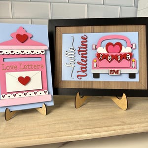 May include: Two Valentine's Day decorations on wooden stands. One is a pink mailbox with a heart and the words "Love Letters." The other is a framed picture of a pink truck with hearts and the words "hello Valentine."
