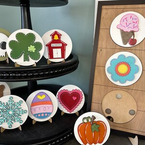 May include: A tiered display stand with various painted wooden ornaments. The ornaments include a skull, a shamrock, a schoolhouse, a turkey, a snowflake, an egg, a heart, a pumpkin, an ice cream cone, and a flower.