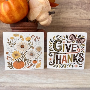 May include: Two square white wooden blocks featuring autumn-themed illustrations. One block displays a pumpkin surrounded by daisies and foliage. The other block features the text "GIVE THANKS" in decorative lettering with a plaid bow graphic.