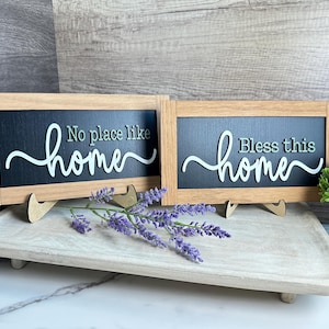 May include: Two framed wooden signs with the phrases "No place like home" and "Bless this home" in white script. The signs are on a wooden tray with lavender sprigs and a small potted plant.