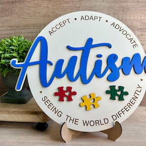 May include: A round wooden sign with the word "Autism" in blue script. The sign includes the phrases "Accept, Adapt, Advocate" and "Seeing the World Differently." Three puzzle pieces in red, yellow, and green are also featured.