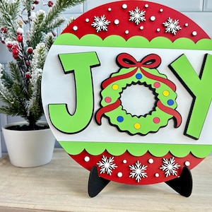 May include: A round, wooden Christmas sign with the word "JOY" in green letters. The "O" is replaced by a wreath with a red bow. The sign has a red and green border with white snowflakes and is displayed on a black stand.