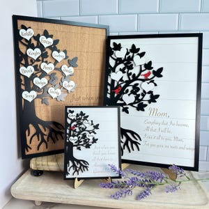 May include: Three framed family tree art pieces. The largest features a black tree with heart-shaped leaves, each bearing a name. The middle piece has a white background with a black tree design and the word "Mom". The smallest piece has a white background.