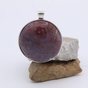 May include: A round silver pendant with a red and purple stone set in the center. The pendant is on a pile of rocks.