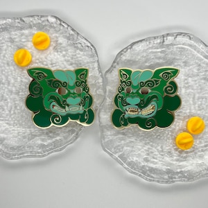 May include: Two green and gold enamel pins depicting a Japanese mythical creature with a fierce expression. The pins are on a clear glass surface with yellow push pins.