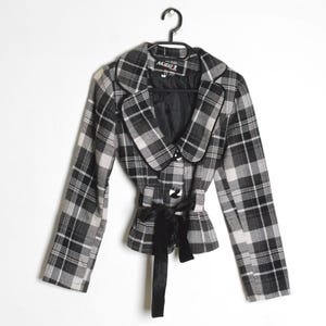 Pode incluir: Um blazer xadrez preto e branco com um cinto de laço de veludo preto. O blazer tem uma gola entalhada e está pendurado em um cabide preto. A etiqueta no blazer diz "Mari Z".