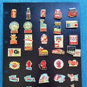 Può includere: Una collezione di 36 spille Coca-Cola con vari design, tra cui i Giochi Olimpici, gli Stati Uniti, Burger King e spille a tema musicale. Le spille sono esposte su uno sfondo nero.
