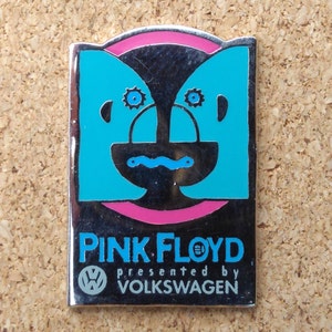 Peut inclure: Une épingle en argent avec un cercle rose et une image bleue et noire d'un visage. Le texte "PINK FLOYD présenté par VOLKSWAGEN" est sous l'image.