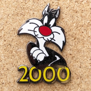 May include: A pin featuring Sylvester the Cat from Looney Tunes. The pin has a black and white design with a red nose and the number "2000" in yellow. The pin is set against a corkboard background.