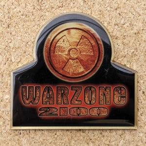 Puede incluir: Un pin negro y dorado con un símbolo radiactivo rojo y naranja y el texto "WARZONE 2100".