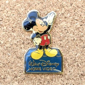 Op de afbeelding: Een gouden pin met Mickey Mouse in een rood-zwart outfit, die met zijn rechterhand zwaait. De pin is op een blauwe achtergrond met de tekst "Walt Disney Home Video".