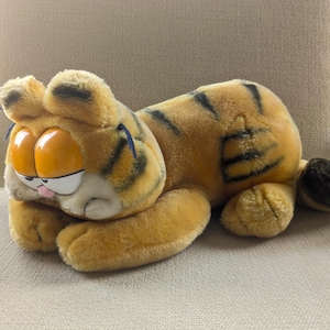 Puede incluir: Un peluche de Garfield en posición acostada. El gato naranja tiene rayas negras, ojos grandes y naranjas y una expresión de mal humor. El juguete está sobre una superficie de color claro.
