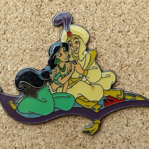 May include: An enamel pin depicting Aladdin and Jasmine on a magic carpet. Aladdin is wearing a white and gold outfit, while Jasmine is wearing a green and blue outfit. The pin is a great addition to any Disney fan's collection.