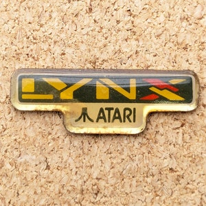Puede incluir: Un pin de logotipo de consola de juegos portátil Atari Lynx dorado y negro. El logotipo presenta el texto "LYNX" en letras amarillas y negras con un diseño gráfico rojo y amarillo. El texto "ATARI" está debajo del logotipo en letras doradas.