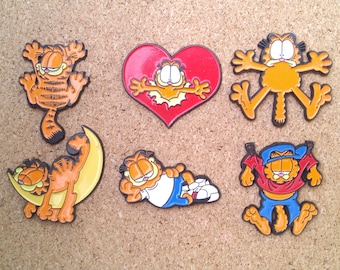 May include: A set of six colorful enamel pins featuring the cartoon character Garfield in various poses. The pins are all different shapes and sizes, and they are all made of metal. The pins are perfect for adding a touch of fun to your clothing, bags, or other accessories.