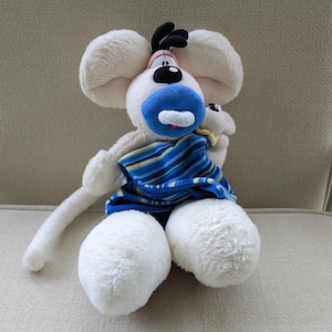 Könnte beinhalten: Ein Stofftier-Maus mit großen weißen Ohren, einer blauen Nase und einem schwarz-rosa Gesicht. Die Maus trägt eine blau-gelb gestreifte Decke. Das Spielzeug sitzt auf einem beigefarbenen Stuhl.