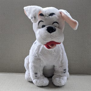 Può includere: Un peluche a forma di cane bianco con macchie nere e bocca rossa seduto su una sedia. Il cane ha grandi occhi neri e orecchie interne rosa. Il giocattolo è fatto di materiale morbido e soffice.