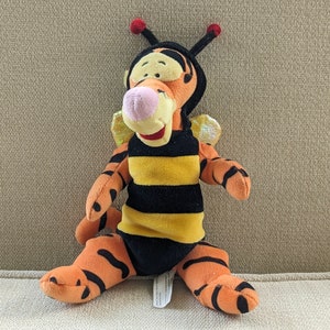May include: A plush toy of Tigger from Winnie the Pooh dressed as a bee. He is wearing a black and yellow striped costume with wings and a red and black antenna.