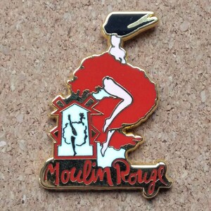 May include: A gold-toned pin featuring a red-skirted can-can dancer with a black hat. The pin includes the words "Moulin Rouge" in a gold script, and a small illustration of the Moulin Rouge building.