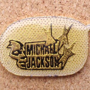 K&ouml;nnte beinhalten: Eine goldfarbene Anstecknadel mit schwarzem Rand und wei&szlig;em Hintergrund. Die Anstecknadel zeigt ein stilisiertes Bild eines Mikrofons und den Text "MICHAEL JACKSON".