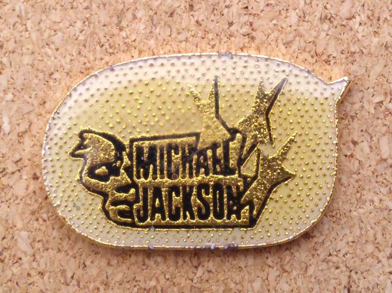 K&ouml;nnte beinhalten: Eine goldfarbene Anstecknadel mit schwarzem Rand und wei&szlig;em Hintergrund. Die Anstecknadel zeigt ein stilisiertes Bild eines Mikrofons und den Text "MICHAEL JACKSON".