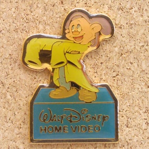 Op de afbeelding: Een gouden pin met een cartoon afbeelding van een dwerg uit de Disney-film Sneeuwwitje en de Zeven Dwergen. De dwerg draagt een geel jasje en staat op een blauwe doos met de tekst "Walt Disney Home Video".