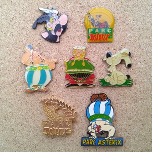 May include: A collection of seven colorful enamel pins featuring characters from the Asterix comic series. The pins depict Asterix, Obelix, Idefix, and other characters from the series. The pins are all in the shape of a circle and have a gold-colored metal backing. The pins are all in good condition and have no visible damage.
