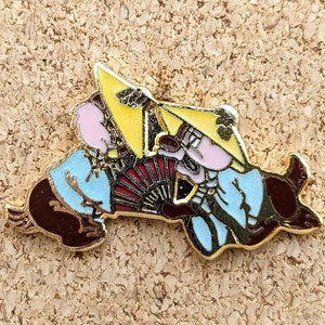 Op de afbeelding: Een gouden speld met een cartoonfiguur dat een gele hoed draagt en een rode en zwarte waaier vasthoudt. Het figuur draagt een blauw-witte gepatroneerde robe.