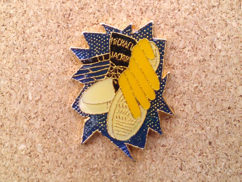 K&ouml;nnte beinhalten: Eine goldfarbene Anstecknadel mit blauem und wei&szlig;em Hintergrund. Die Nadel zeigt einen gelben Schuh mit dem Text "MICHAEL JACKSON" an der Seite.