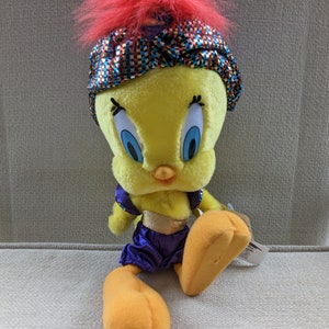 Op de afbeelding: Een gele pluche Tweety Bird die een paars-gouden kostuum draagt met een rood-gouden tulband. Het pluche speelgoed zit op een lichtbruin oppervlak.