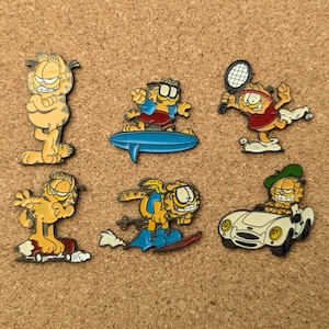 Puede incluir: Colección de seis pines del personaje Garfield en varias poses. Un pin muestra a Garfield de pie con los brazos cruzados, mientras que otros lo representan surfeando, jugando al tenis, esquiando y conduciendo un coche blanco. Los pines están sobre un fondo de corcho.