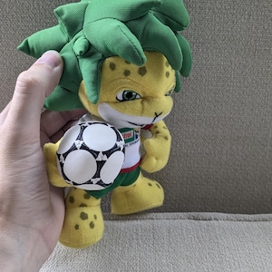 May include: A plush toy mascot with a yellow body, green spiky hair, and a soccer ball. The mascot has a friendly expression and is wearing a white shirt with a logo. The toy is held by a hand.