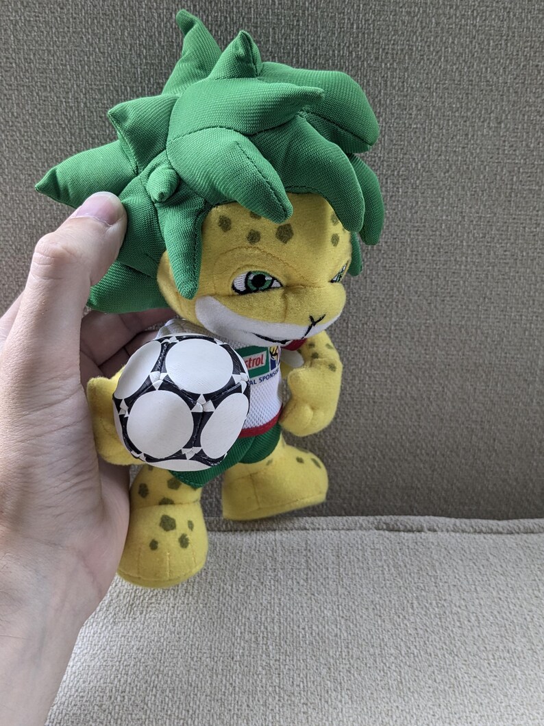 May include: A plush toy mascot with a yellow body, green spiky hair, and a soccer ball. The mascot has a friendly expression and is wearing a white shirt with a logo. The toy is held by a hand.