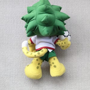 May include: A plush toy mascot with a green spiky head, white shirt, and green shorts. The mascot has yellow spotted feet and tail, and is holding a soccer ball. The toy is designed with a playful and friendly appearance.