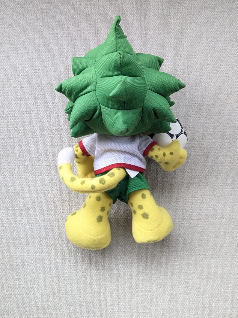 May include: A plush toy mascot with a green spiky head, white shirt, and green shorts. The mascot has yellow spotted feet and tail, and is holding a soccer ball. The toy is designed with a playful and friendly appearance.