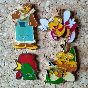 Op de afbeelding: Vier emaillepins met cartoonfiguren. Een bruine beer die een overall en een rood bandana draagt met het woord "Chocos" erop. Een gele en zwarte bij met een rood-witte sjaal. Een groene haan met een rode kam. Een gele en bruine bij met een bril en een groen blad.