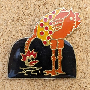 Puede incluir: Un pin de esmalte colorido con una avestruz naranja y amarilla con lunares rojos, inclinándose para mirar a un pájaro rojo y amarillo más pequeño sobre un fondo negro.