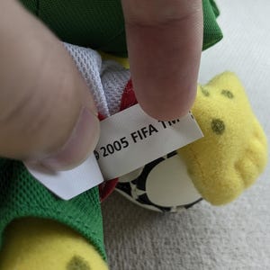 May include: A close-up of a plush toy with a tag that reads "2005 FIFA TM." The toy has yellow, green, and white elements. The tag is white with black text. The toy's design includes a soccer ball.