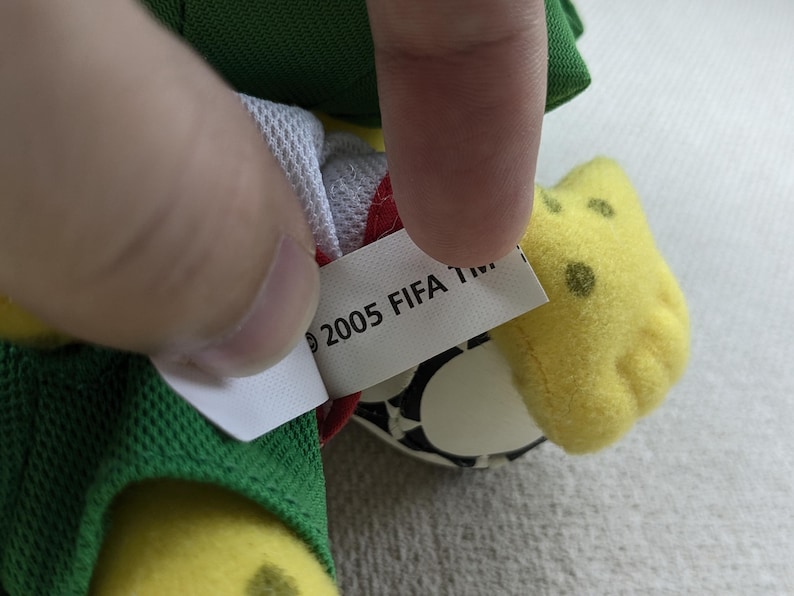 May include: A close-up of a plush toy with a tag that reads "2005 FIFA TM." The toy has yellow, green, and white elements. The tag is white with black text. The toy's design includes a soccer ball.