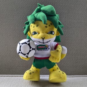 Op de afbeelding: Een pluche speelgoed van een gele jaguar-achtige mascotte met groen haar, gekleed in een wit en rood shirt, groene shorts en gele schoenen. De mascotte houdt een zwart-witte voetbal vast. Het shirt heeft een sponsorlogo.