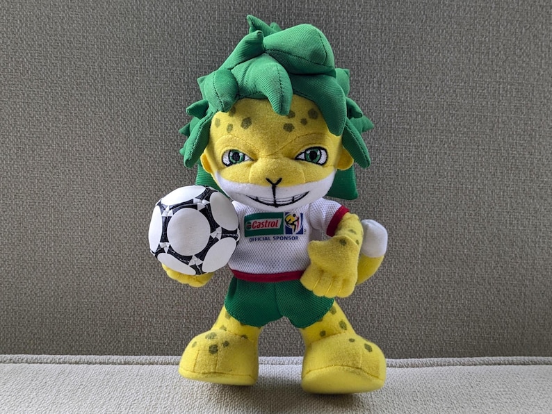 May include: A plush toy of a yellow jaguar-like mascot with green hair, wearing a white and red shirt, green shorts, and yellow shoes. The mascot is holding a black and white soccer ball. The shirt has a sponsor logo.